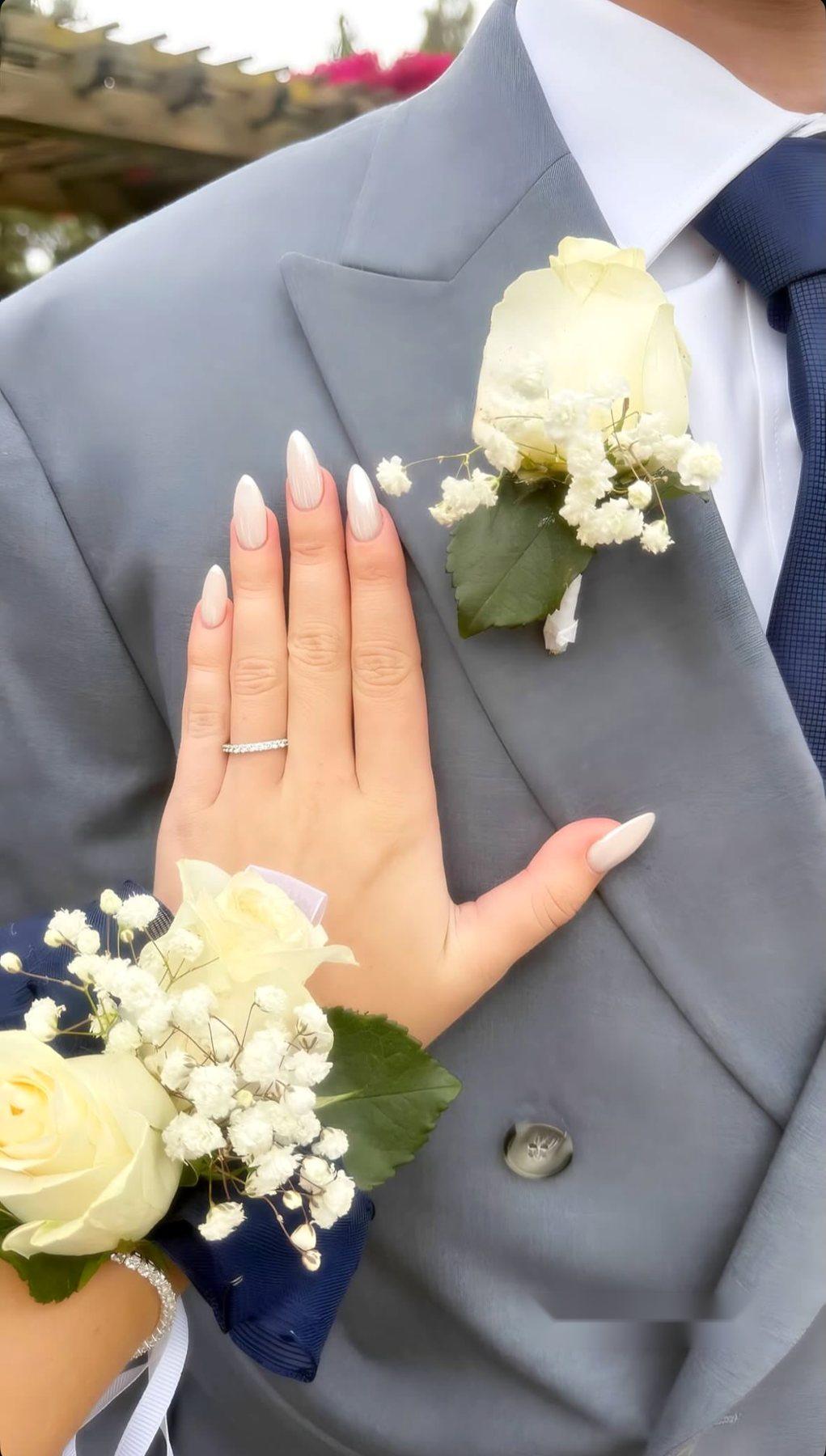 A white rose corsage on a navy ribbon, worn on the wrist against a gray suit with a white rose boutonnière