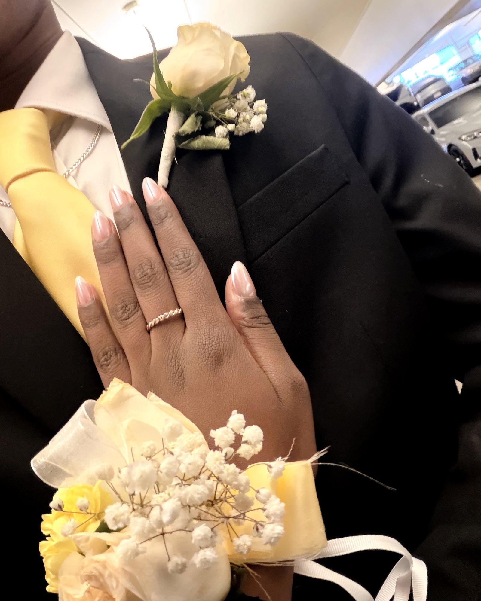 A yellow and white rose corsage on the wrist against a black suit with a yellow tie and a white rose boutonnière
